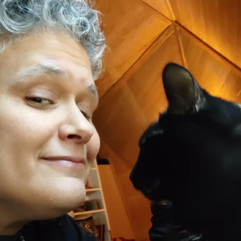 Vas Vas, a silver-haired person of mixed ethnicity is hanging out with a black cat.